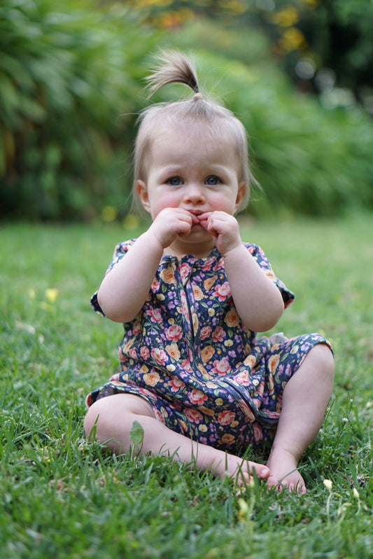 A toddler girl sitting on green grass in a garden, wearing a short-sleeve floral romper from Little Acres Co.